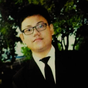 Ibu Diah's son, a picture of him in front of many trees. He is wearing a suit and tie and has glasses on.